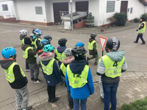Foto der Klasse. Die Kinder stehen in gelben Westen auf der Strße und bekommen Erklärungen zu den Verkehrsschildern.